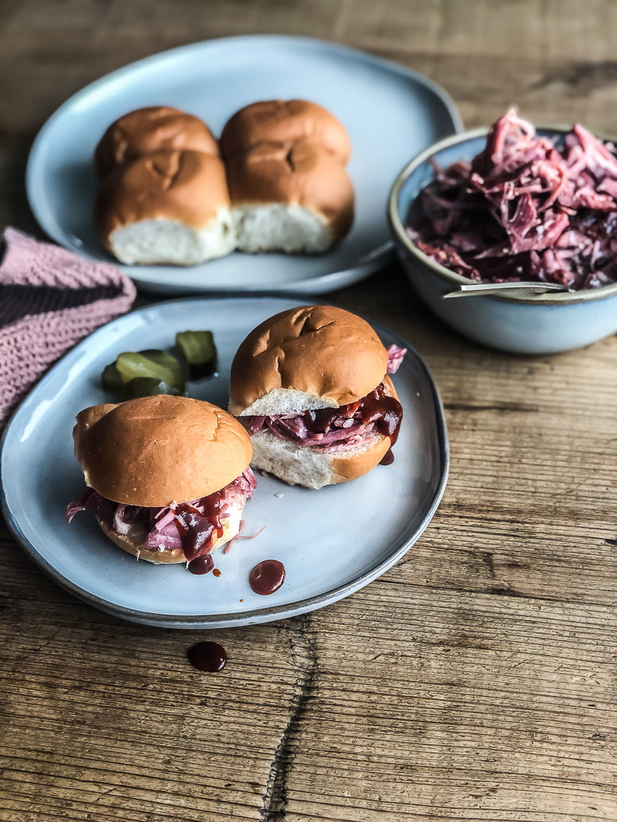 pulled pork sandwiches on plates on wooden table with pickles and barbecue sauce